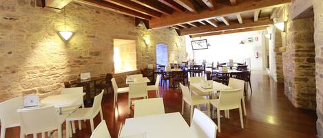 A spacious dining area with white tables and chairs, stone walls, and wooden beams.