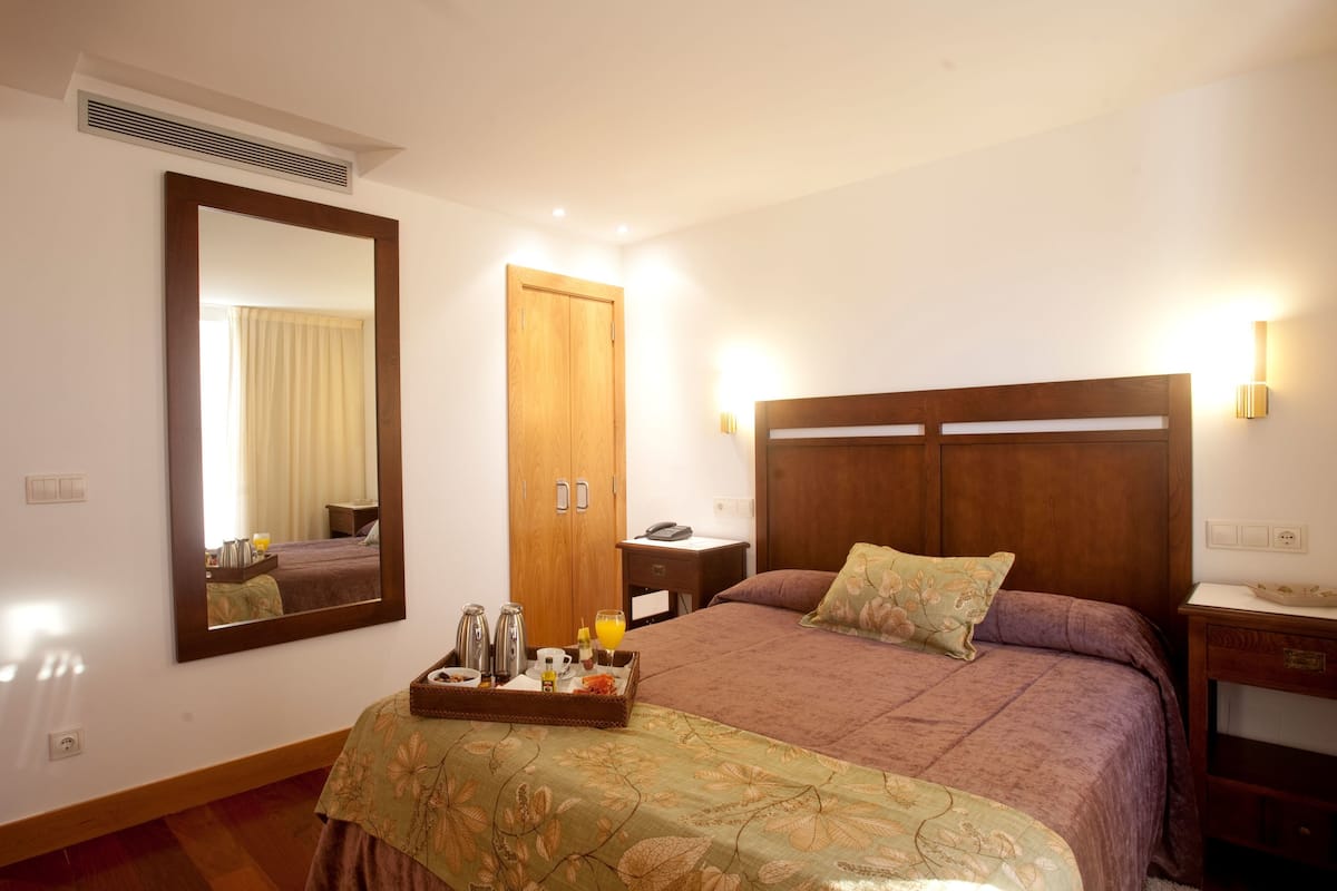 Ein Hotelzimmer mit einem großen Bett, einem hölzernen Kopfteil, einem Nachttisch, einem Spiegel und einer Ablage mit Frühstücksgegenständen.