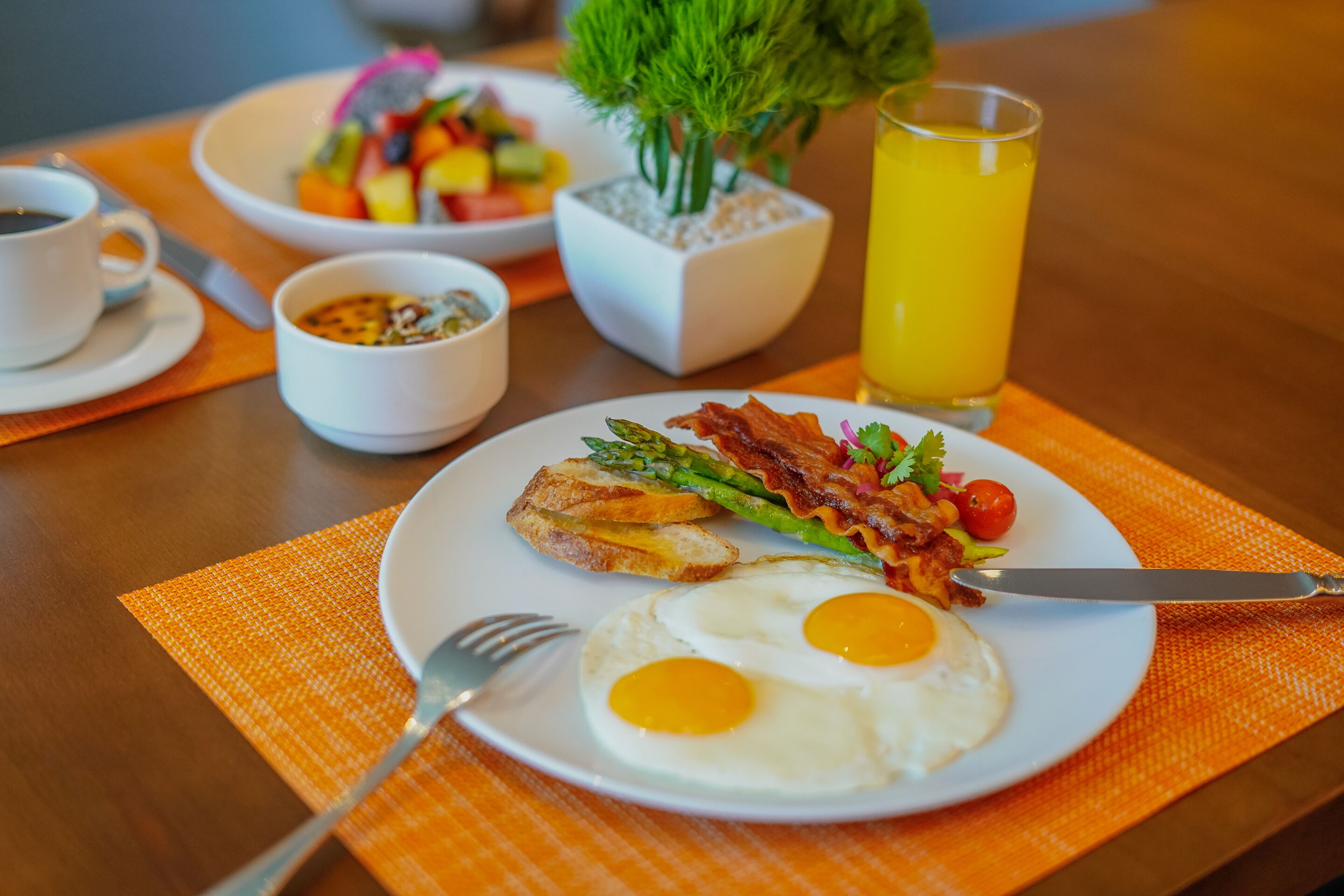 A breakfast plate with eggs, bacon, and toast, a bowl of fruit, a cup of coffee, and a glass of orange juice on a wooden table.