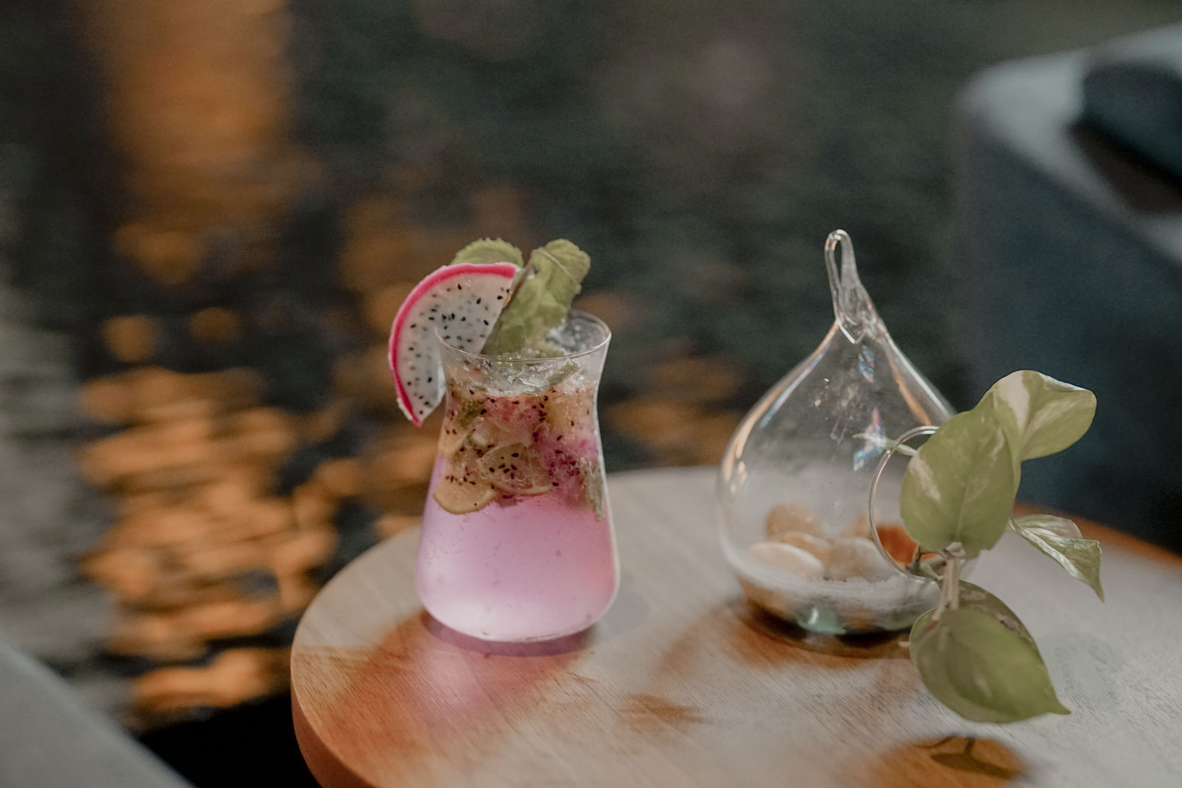 Um cocktail com uma fatia de pitaya e folhas de hortelã num copo, colocado sobre uma mesa de madeira ao lado de um pequeno copo com uma planta.