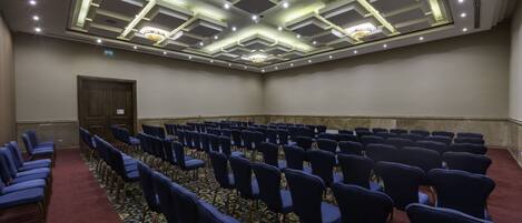 Un salón de conferencias con filas de sillas azules, alfombra estampada y un diseño de techo moderno con iluminación empotrada.