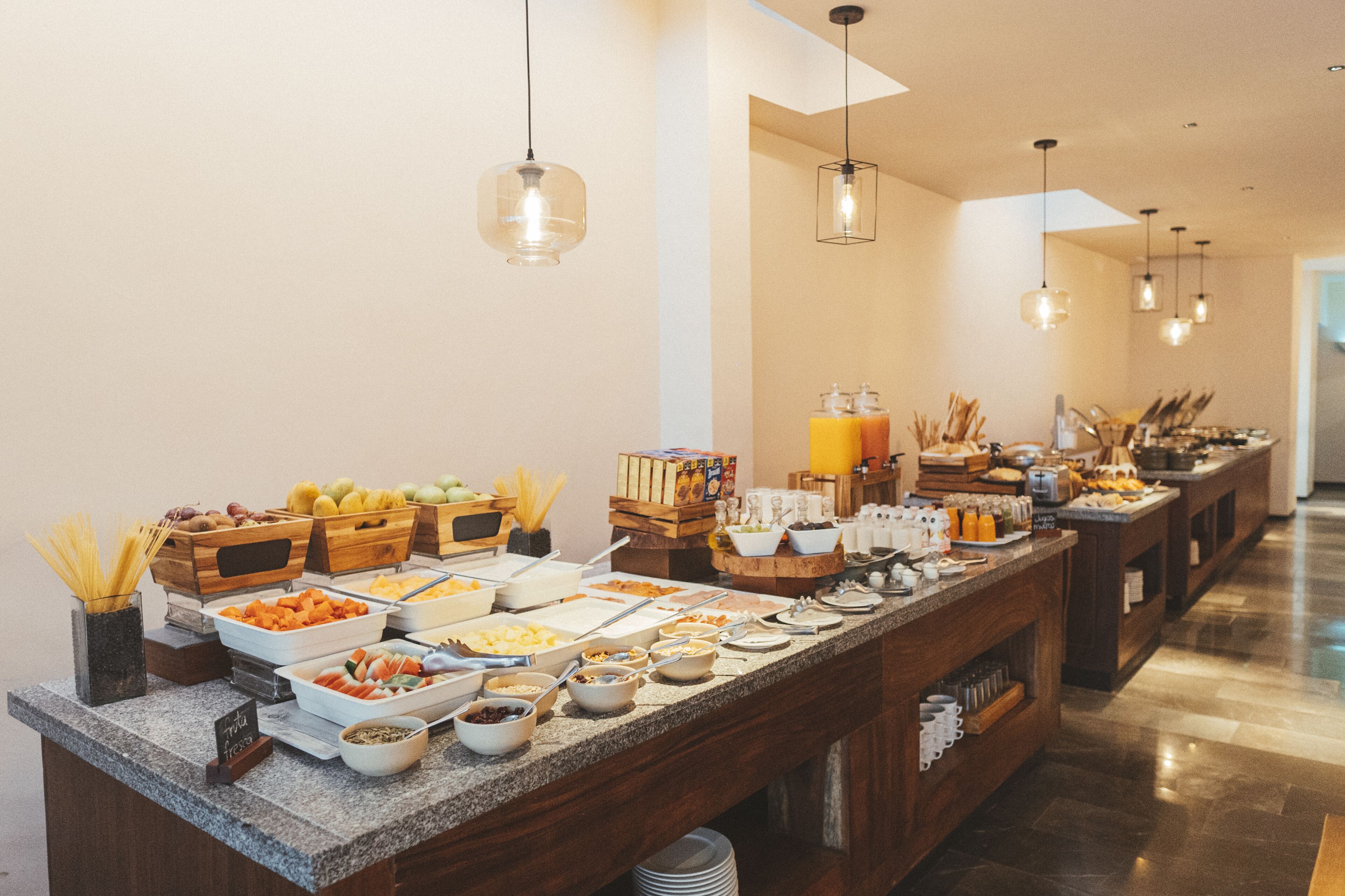 A buffet setup with various food items and beverages, wooden furniture, and hanging pendant lights.