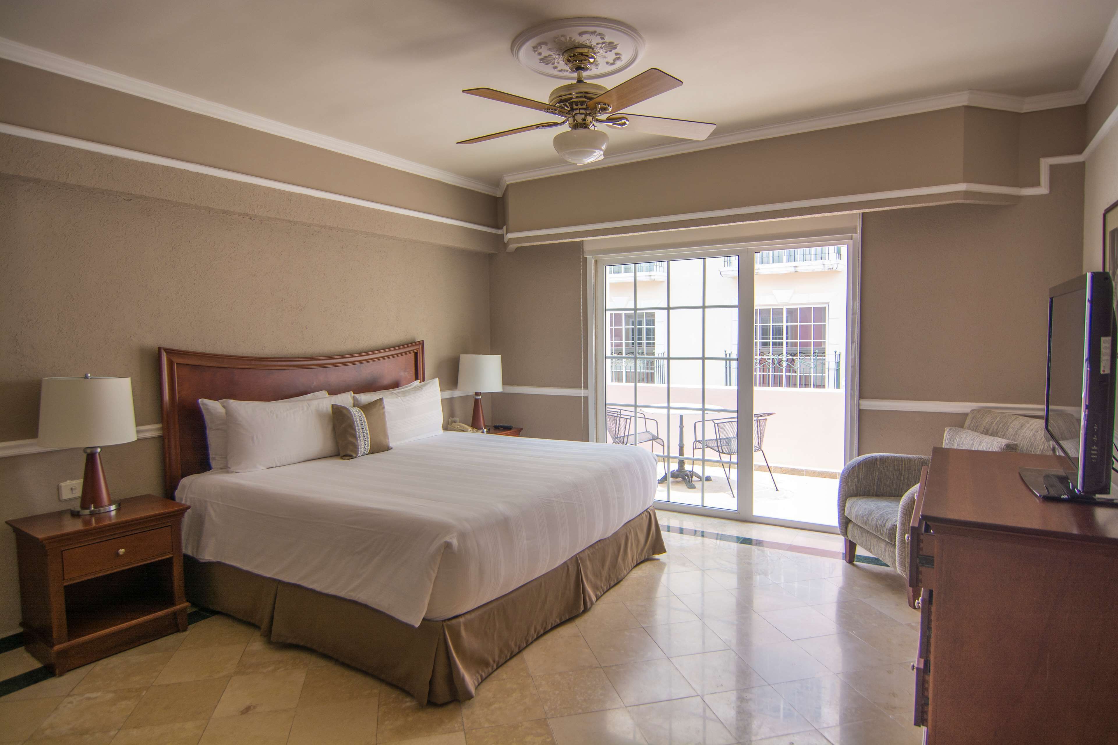 A hotel room with a large bed, two bedside lamps, a ceiling fan, a TV, and a balcony with a chair.