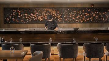 A restaurant with a chef preparing food, dark wooden tables, and chairs, and a large abstract wall art.