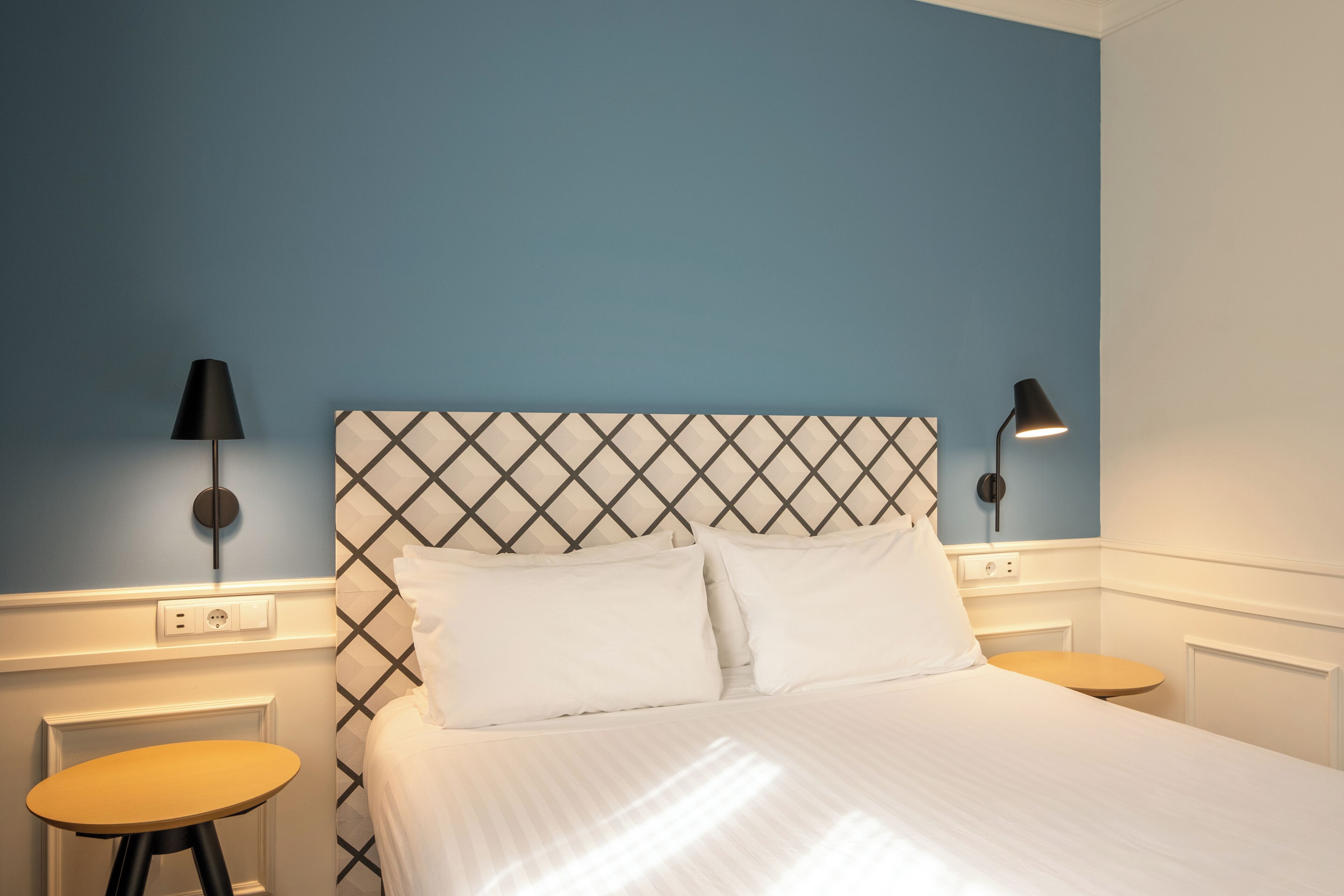 Un lit bien fait, avec du linge de lit blanc, une tête de lit à motifs losangés et deux tables de chevet avec des lampes.