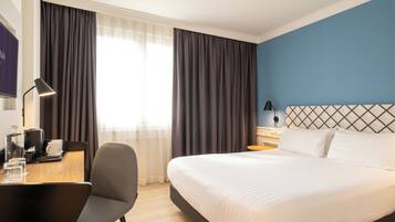 A hotel room with a large bed, a desk with a chair, a lamp, and a view of the outdoors through the window.