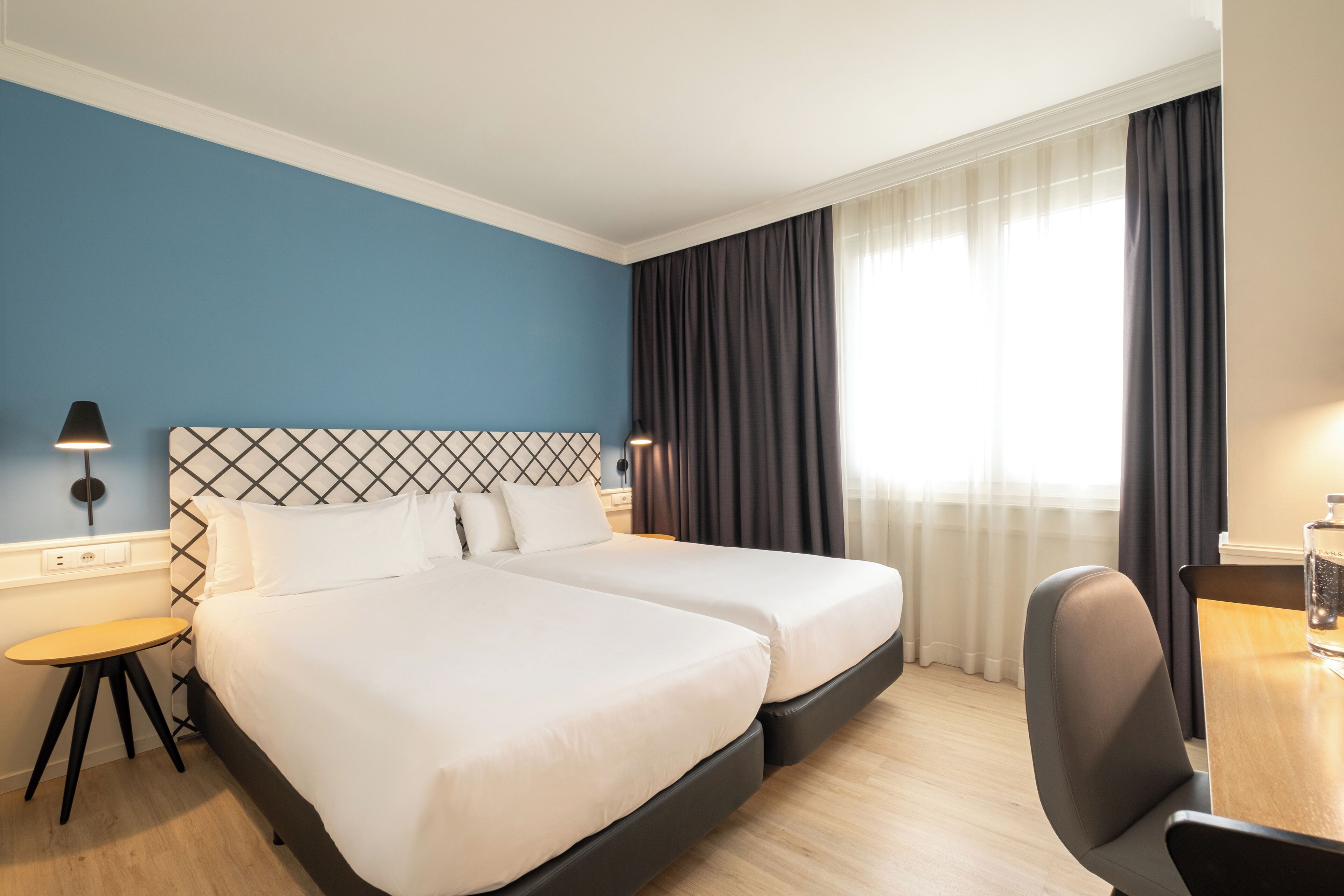 Une chambre d’hôtel avec deux lits, un bureau, une chaise, une lampe et une fenêtre avec des rideaux.