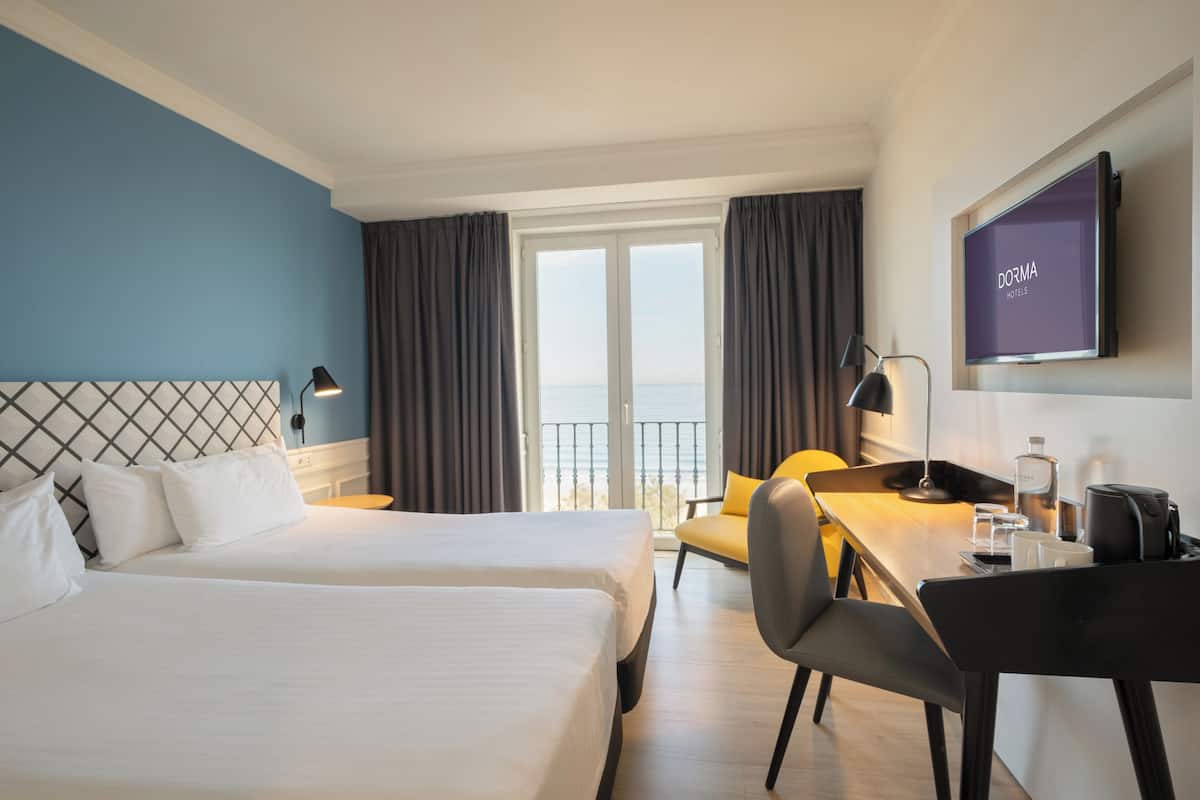 A hotel room with two beds, a desk, a chair, a TV, and a view of the sea.