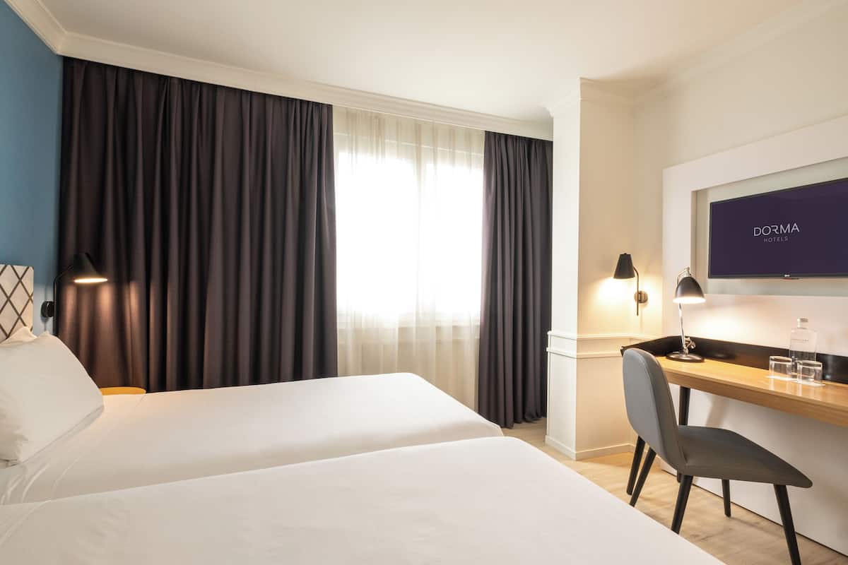 A hotel room with two beds, a desk, a chair, a TV, and a window with curtains.