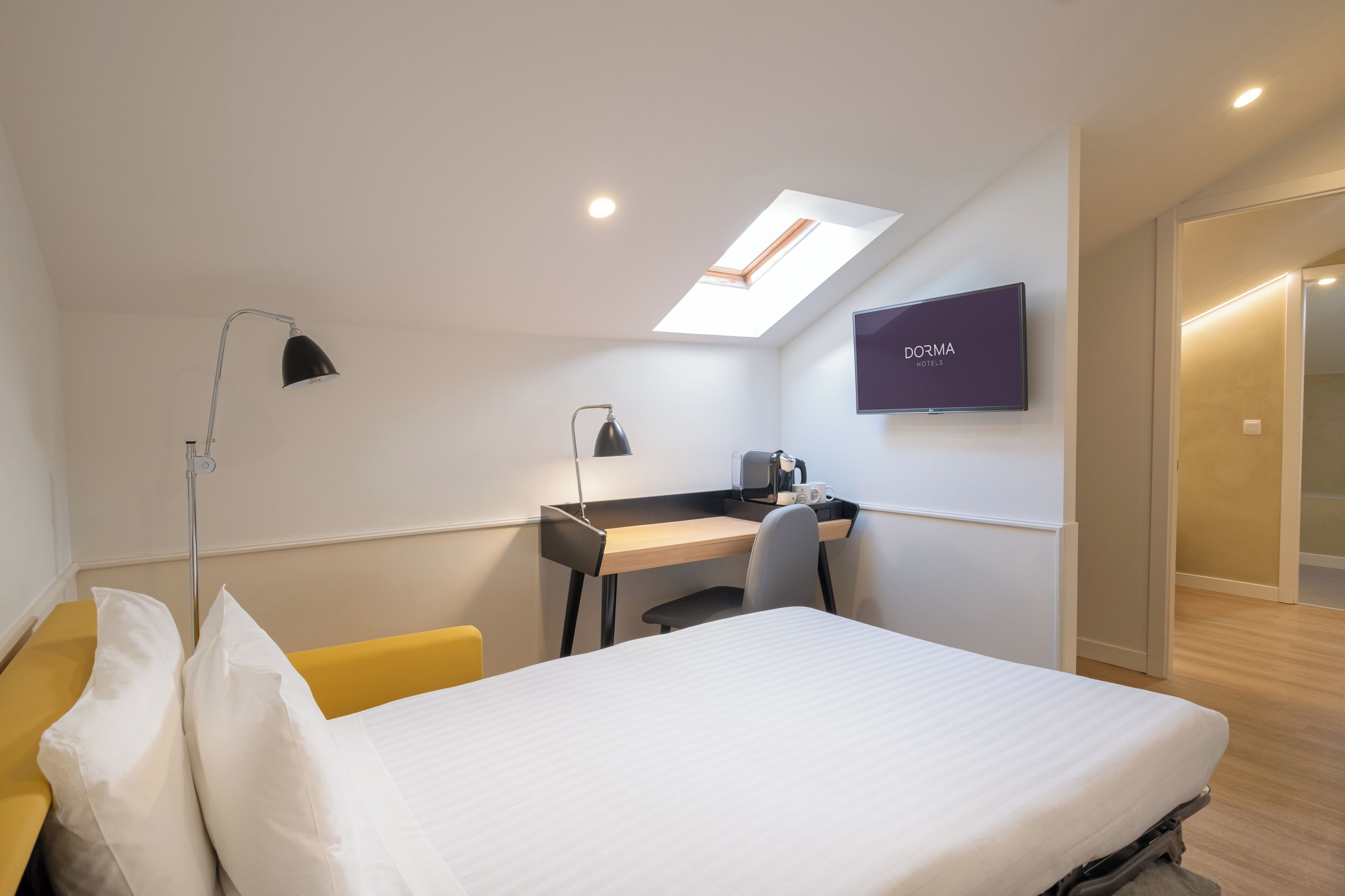 A modern hotel room with a bed, desk, and chair, a TV mounted on the wall, and a skylight above the desk.