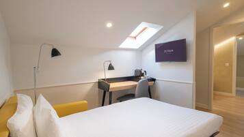 A modern hotel room with a bed, desk, and chair, a TV mounted on the wall, and a skylight above the desk.