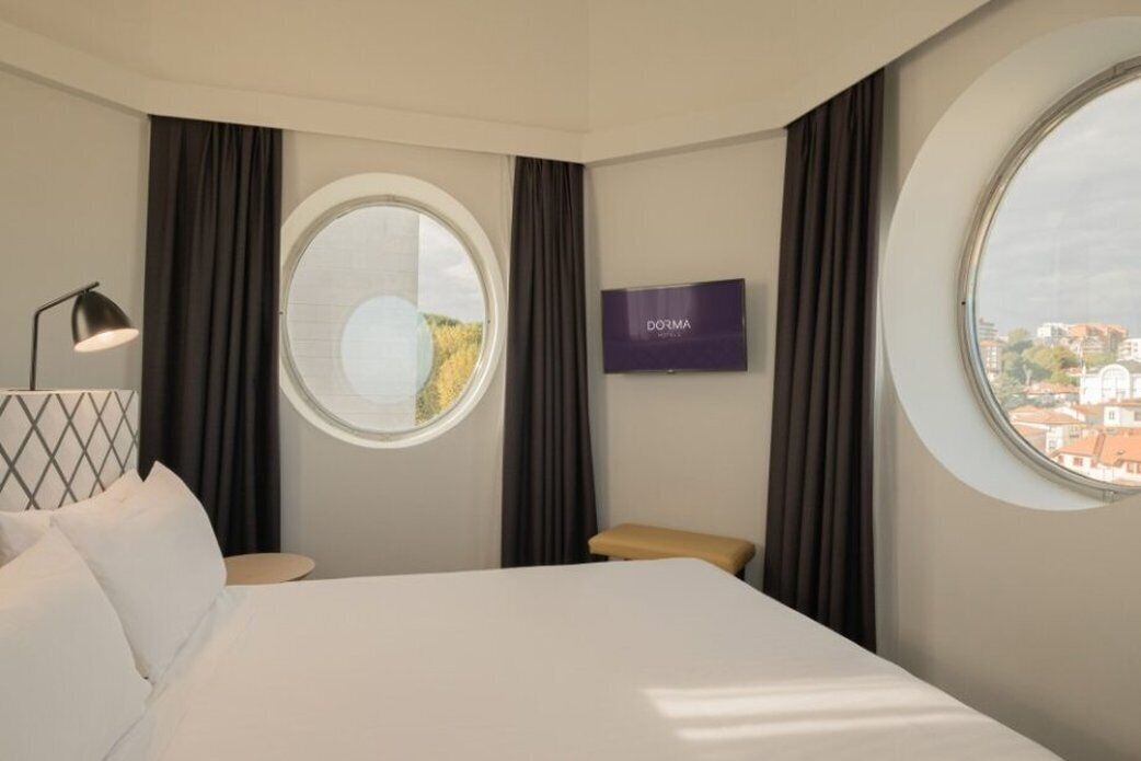 Habitación de hotel con cama, televisión, ventana redonda y vista de la ciudad.