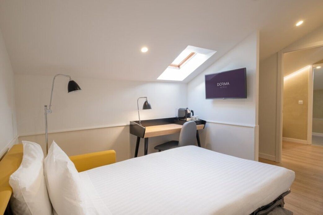 A hotel room with a bed, a desk, a chair, a TV, and a skylight.