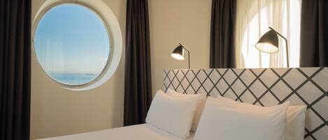 A hotel room with a large bed, a round window, and a view of the sea.