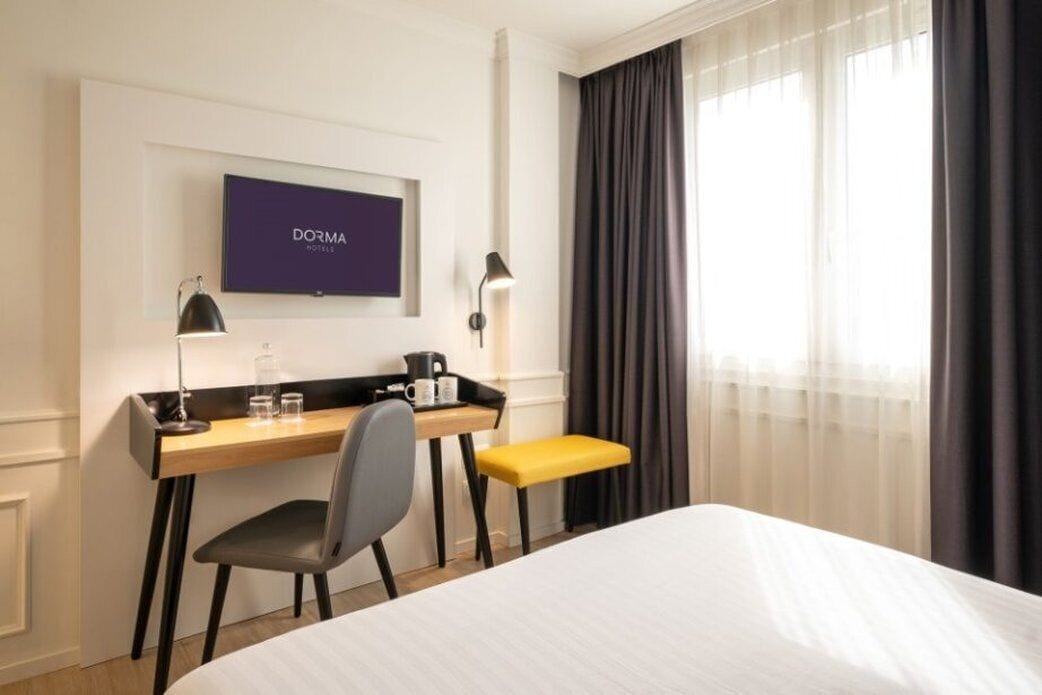 A hotel room with a bed, a desk with a chair, a small table with a bench, a TV mounted on the wall, and a window with curtains.