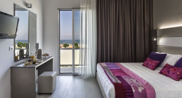 A modern hotel room with a balcony, a bed with decorative pillows, a flat-screen TV, and a breakfast bar with a coffee maker. - Hotel Boemia (Riccione)