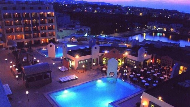 Zona de piscina de hotel con múltiples sillas de descanso y sombrillas, rodeada de edificios con ventanas iluminadas.