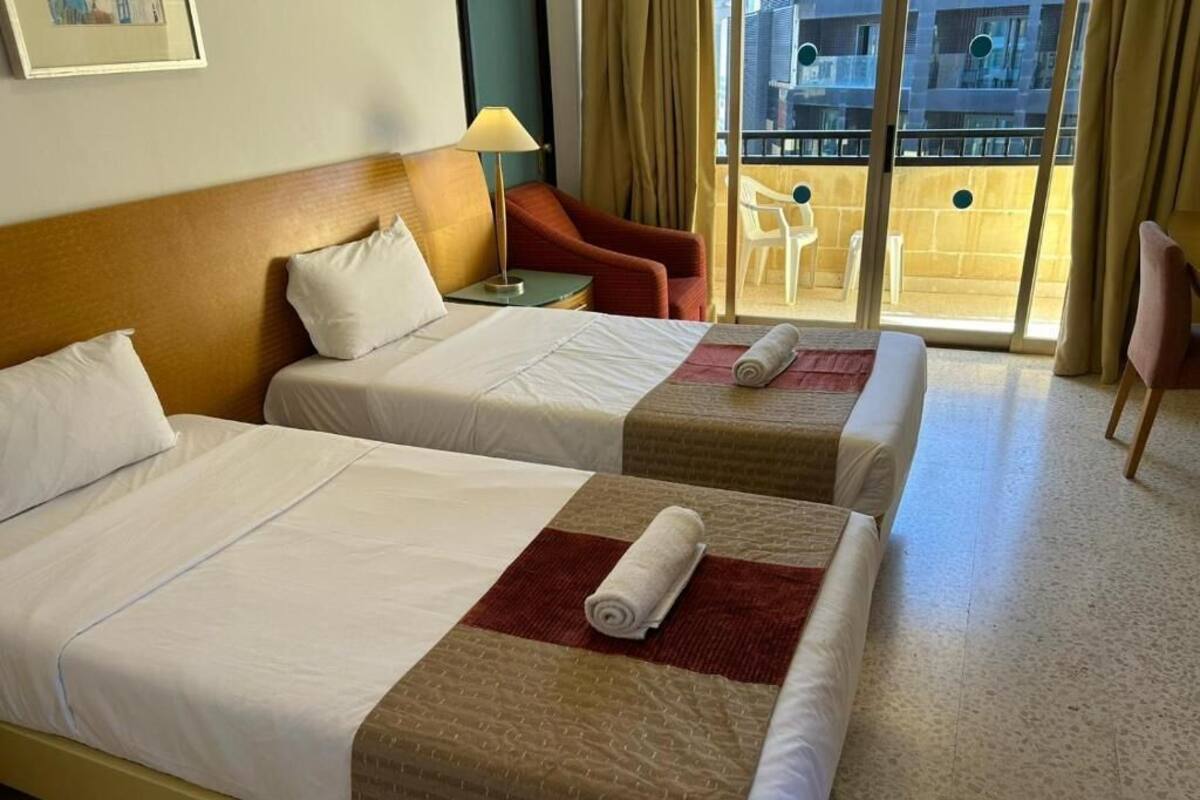 Habitación de hotel con dos camas, un sillón rojo, una mesita y un balcón con vistas a la ciudad.