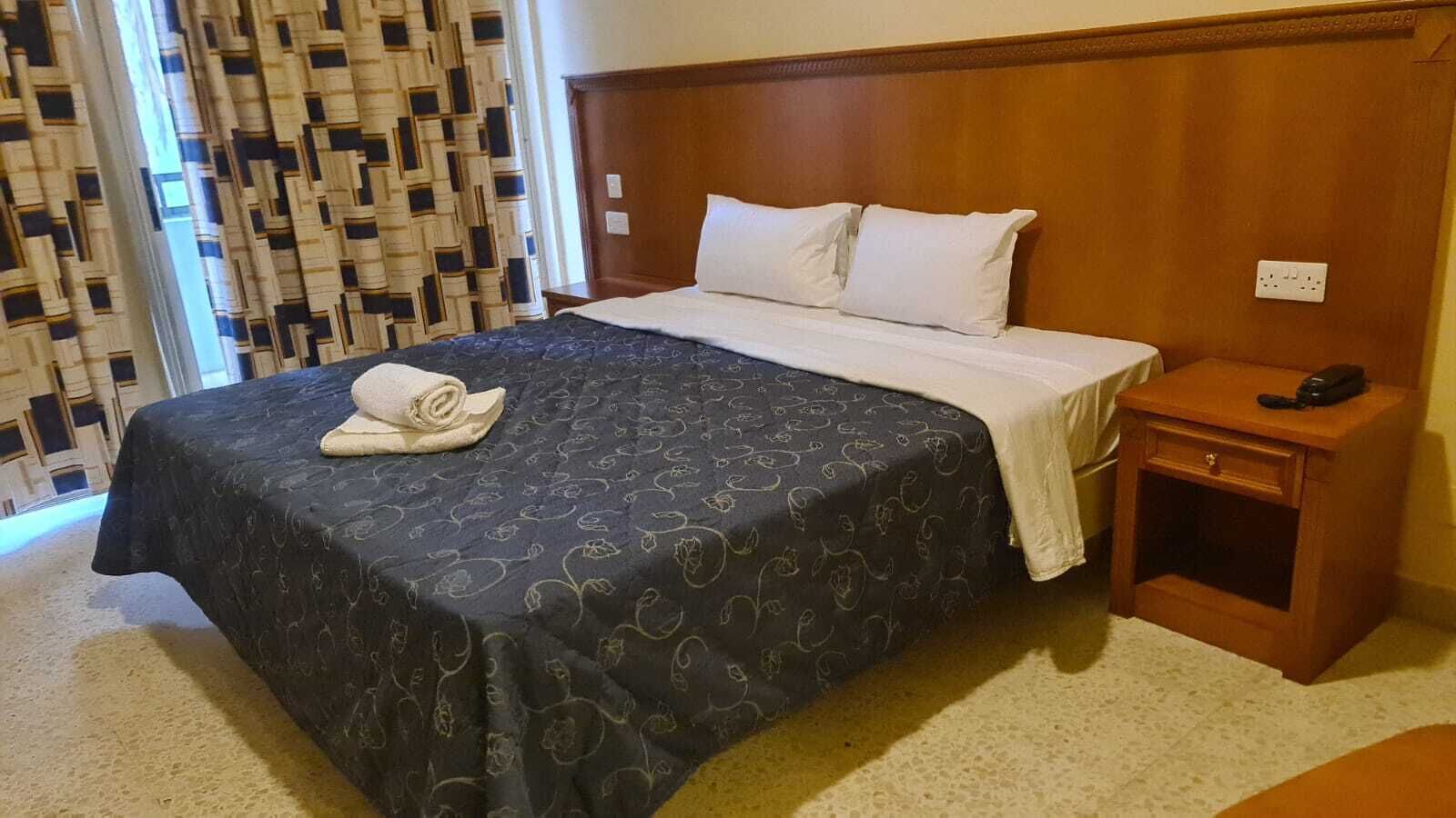 Una cama bien tendida con una colcha oscura estampada y almohadas blancas, una mesita de noche de madera con un teléfono y una cortina con estampado al fondo.