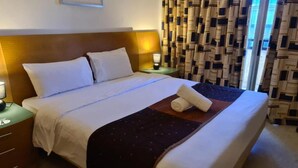 Una cama bien hecha con sábanas blancas, un plaid oscuro estampado y toallas enrolladas sobre un cabecero de madera.