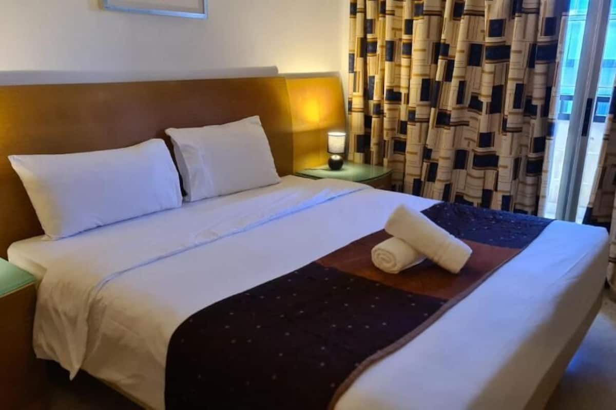 Una cama bien hecha con sábanas blancas, un plaid oscuro estampado y toallas enrolladas sobre un cabecero de madera.