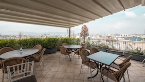 A covered outdoor seating area with tables and chairs, overlooking a cityscape. - Golden City Hotel (Athens)