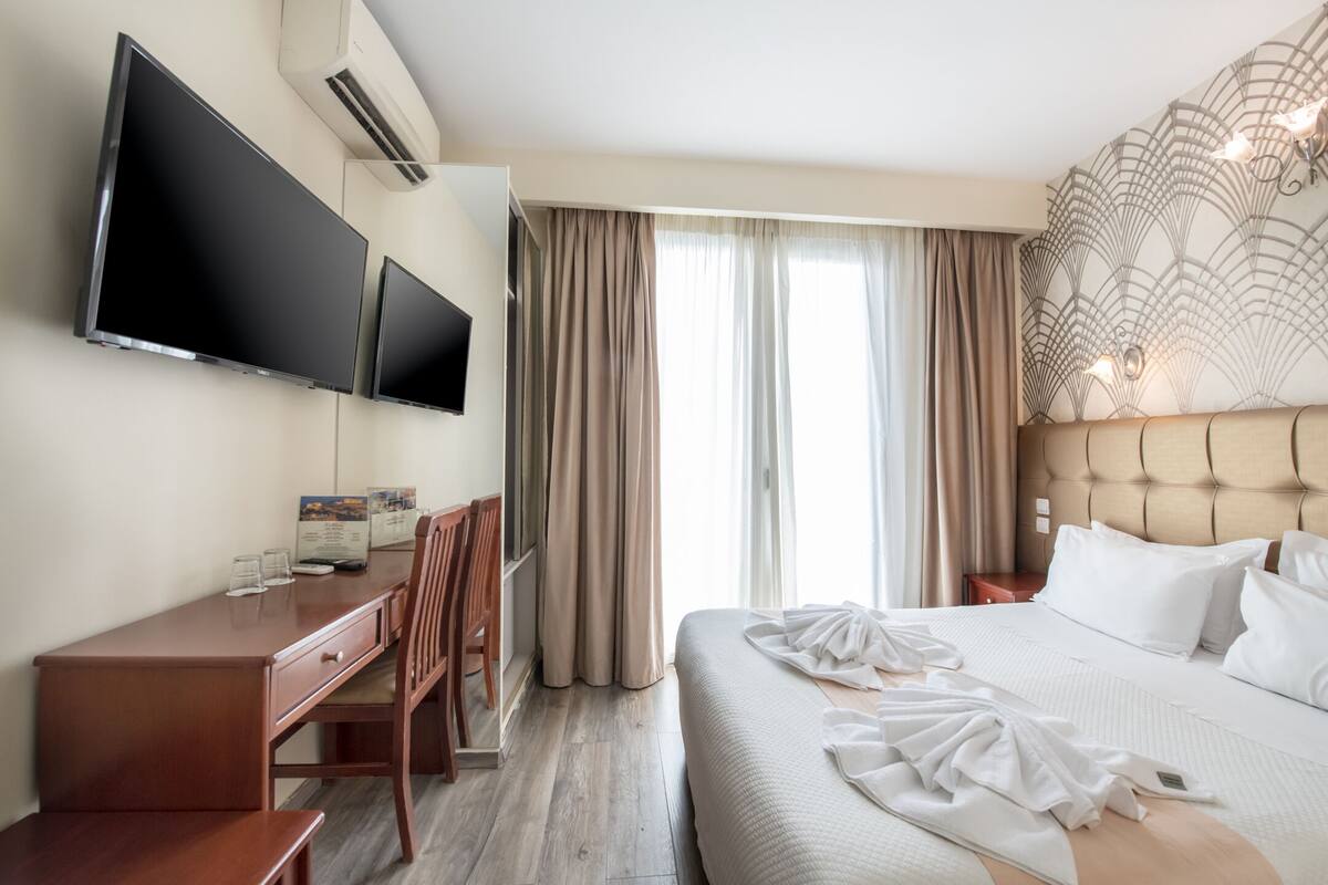 Hotellihuone, jossa on sänky, työpöytä, kaksi televisiota ja ikkuna, jossa on verhot.