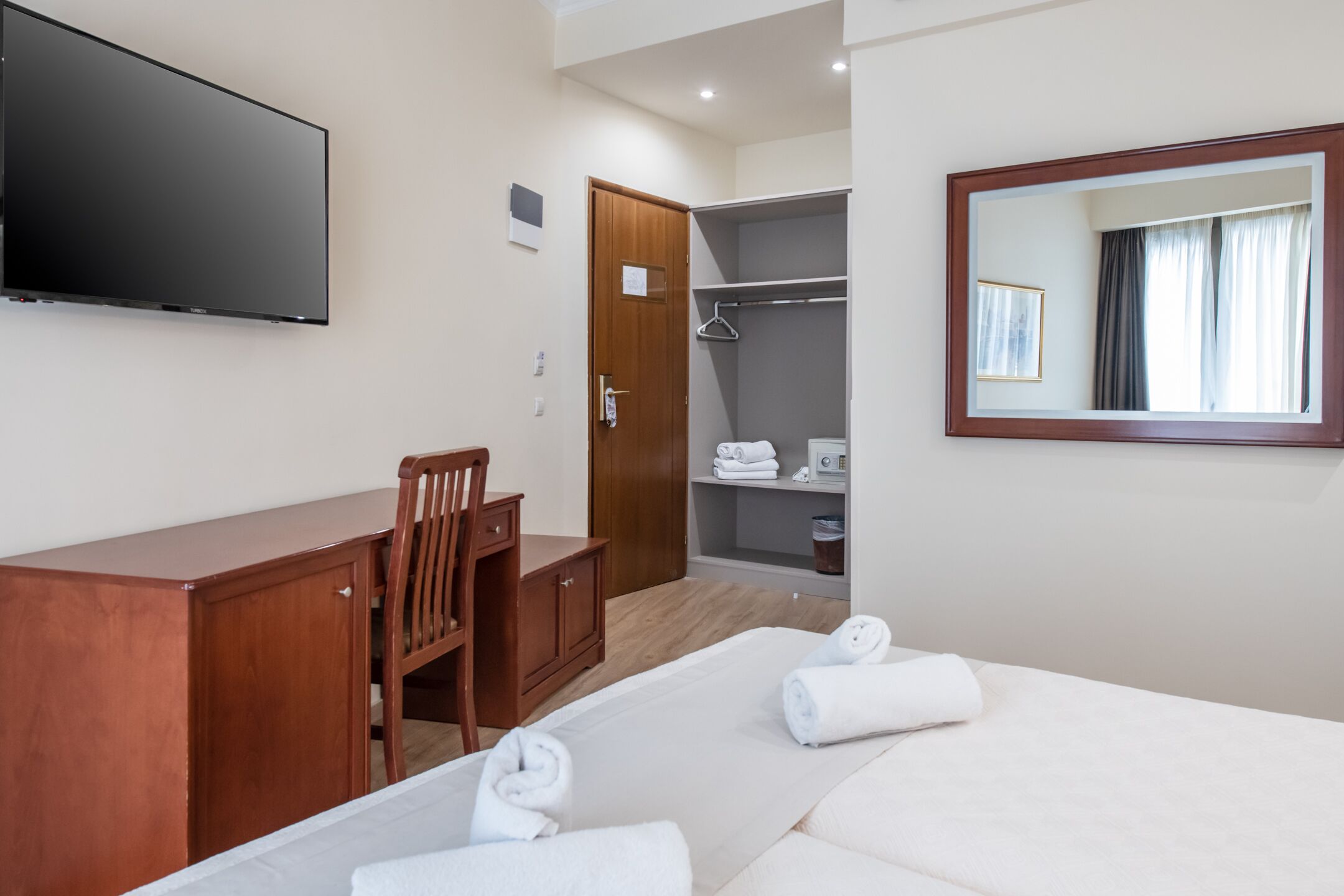 A hotel room with a bed, a desk, a chair, a wardrobe, a television, and a mirror.