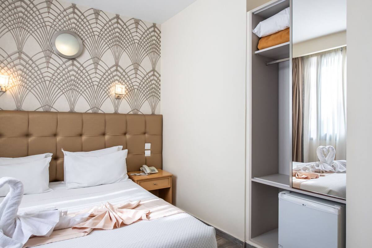 Hotellihuone, jossa on napitettulla päädyllä varustettu sänky, valkoisilla lakanoiden peittämä sänky, yöpöytä lampulla, peilillä varustettu vaatekaappi ja pieni valkoinen jääkaappi.