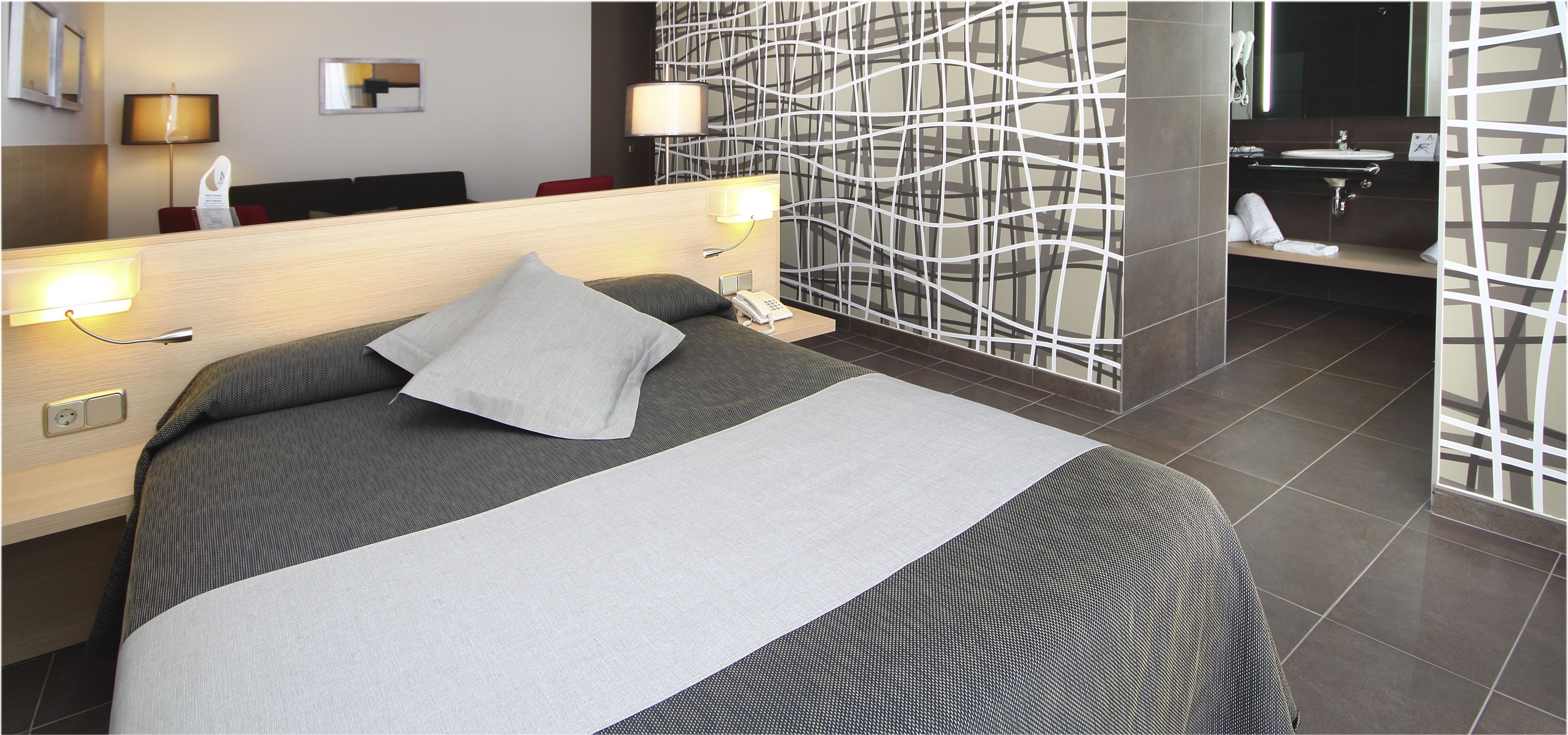 Ein modernes Hotelzimmer mit einem großen Bett, Nachttischlampen, einem Schreibtisch und einem Badezimmer mit modernem Waschbecken und Spiegel.