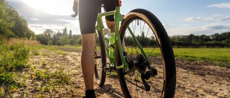 A person standing next to a bicycle on a dirt path with greenery in the background.