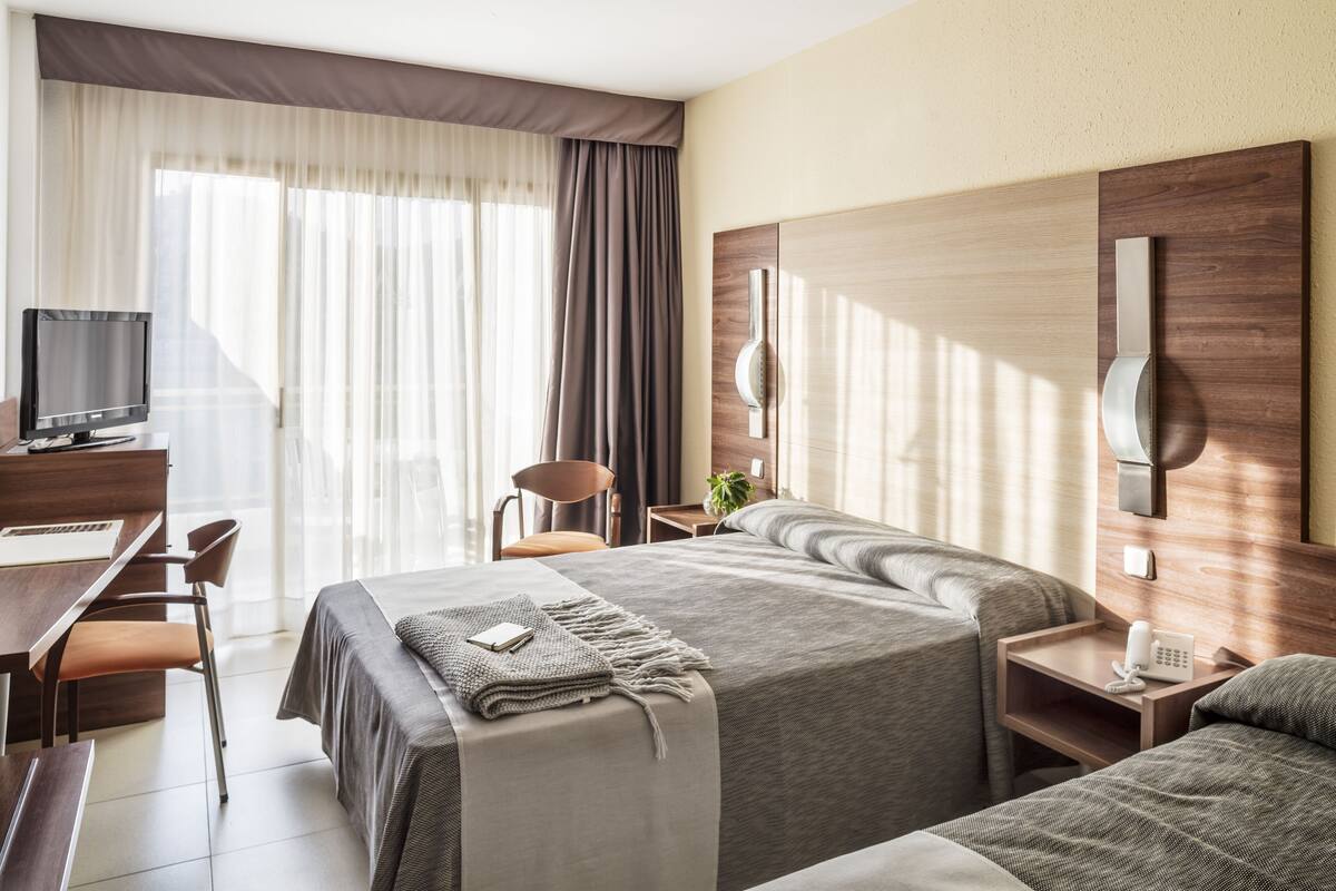 Habitación de hotel con dos camas, un escritorio con computadora, televisor y mesita de noche con teléfono.