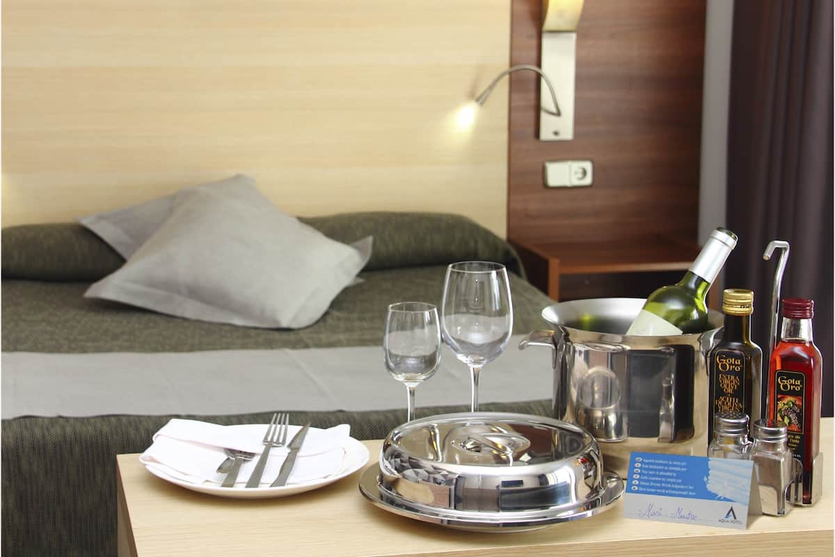 Habitación de hotel impecable con una cama, una mesita de noche con un bandeja de comida, copas de vino y botellas de agua y vino.