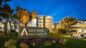 A hotel sign with a palm tree and flowers in the foreground, and a building with balconies in the background. - AQUA Hotel Aquamarina & Spa (Santa Susanna)