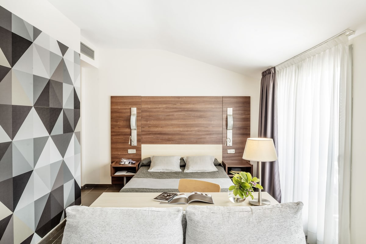 Habitación de hotel moderna con pared de diseño geométrico, cabecera de madera, cama con ropa de cama gris, mesita de noche con lámpara y una planta pequeña.