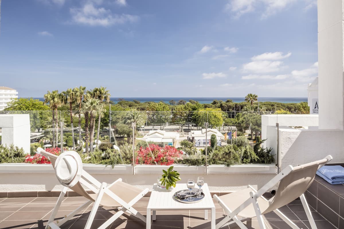 Un balcón con dos sillones blancos, una mesita con una planta en maceta y una bandeja con un vaso de agua. La vista muestra un paisaje costero con palmeras y edificios.