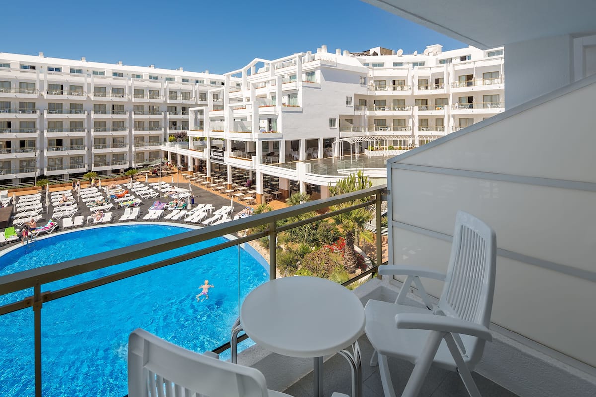 Un balcón de hotel con piscina, sillas blancas y vistas al área de la piscina.