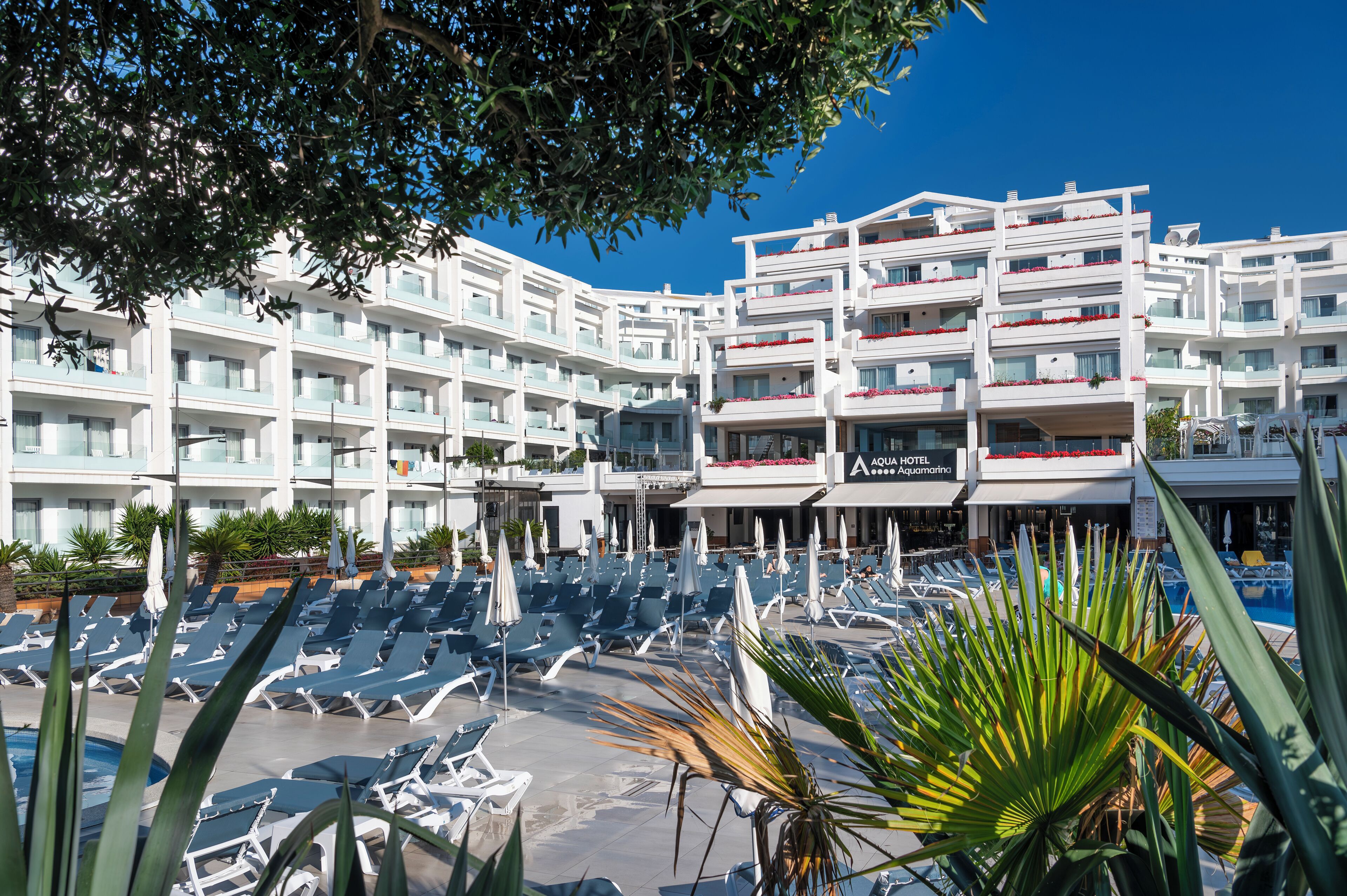 Ein Hotel mit mehreren Balkonen, einem Poolbereich inklusive Liegestühlen und einem strahlend blauen Himmel.