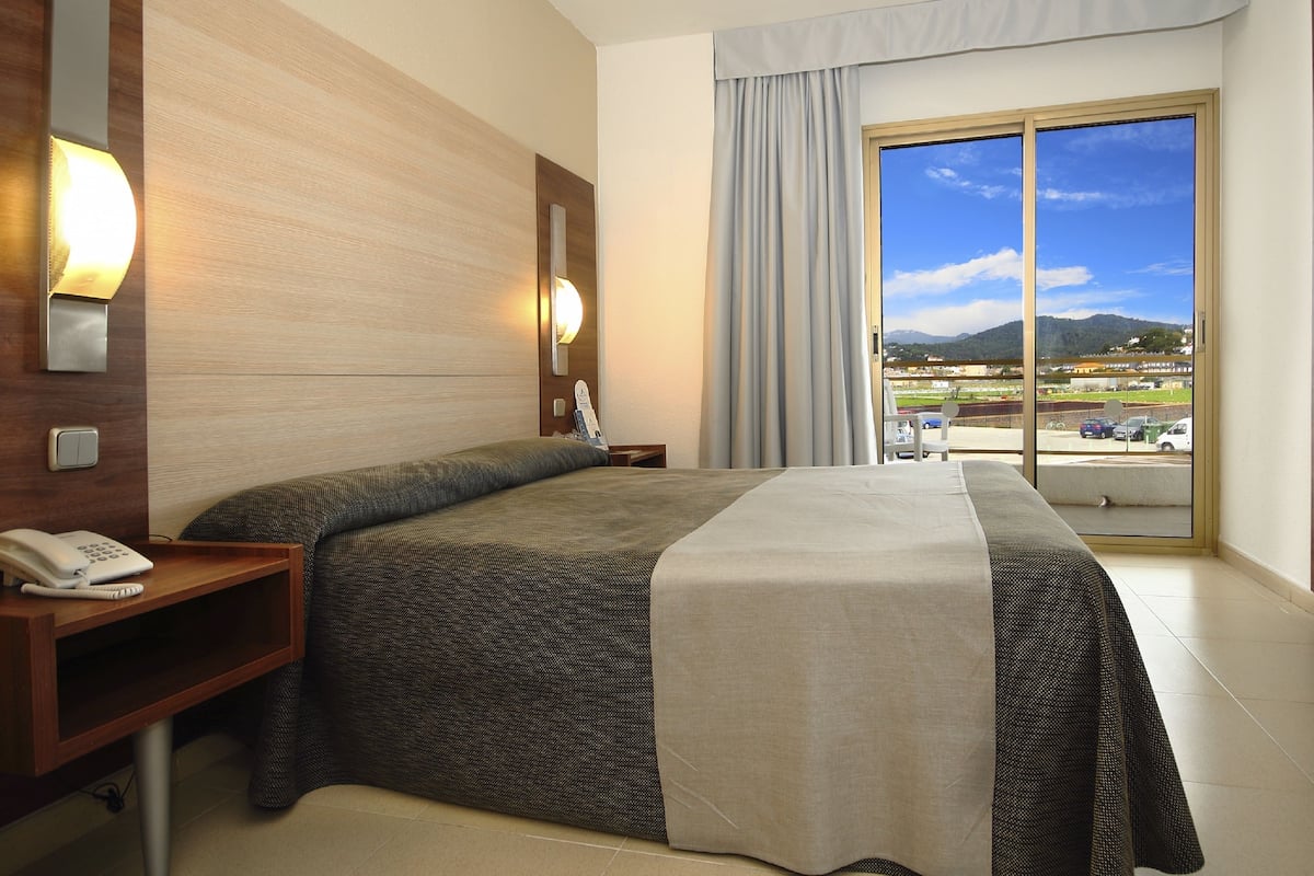 Habitación de hotel con cama, teléfono en la mesita de noche, mesita auxiliar y ventana con vista a un estacionamiento y montañas.