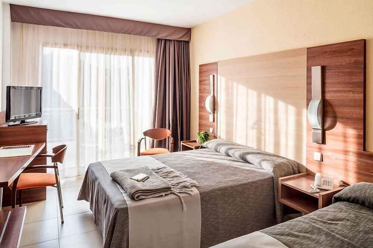 A hotel room with a bed, a desk, a chair, a TV, and a bedside table with a phone.