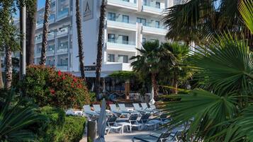 Un hotel con varios pisos, palmeras y un área de piscina con tumbonas.