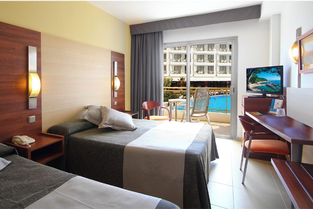 A hotel room with two beds, a desk, a TV, and a balcony with a pool view.