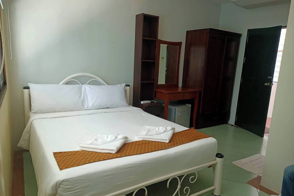 A neatly made bed with white linens and two folded towels, a wooden wardrobe, a small table with a mirror, and a green door.
