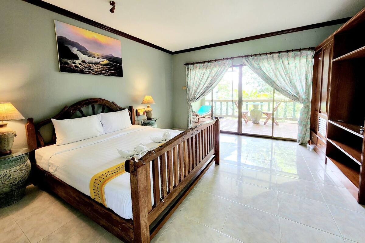 A hotel room with a large bed, a painting of a seascape, a balcony with a view, and a wooden wardrobe.