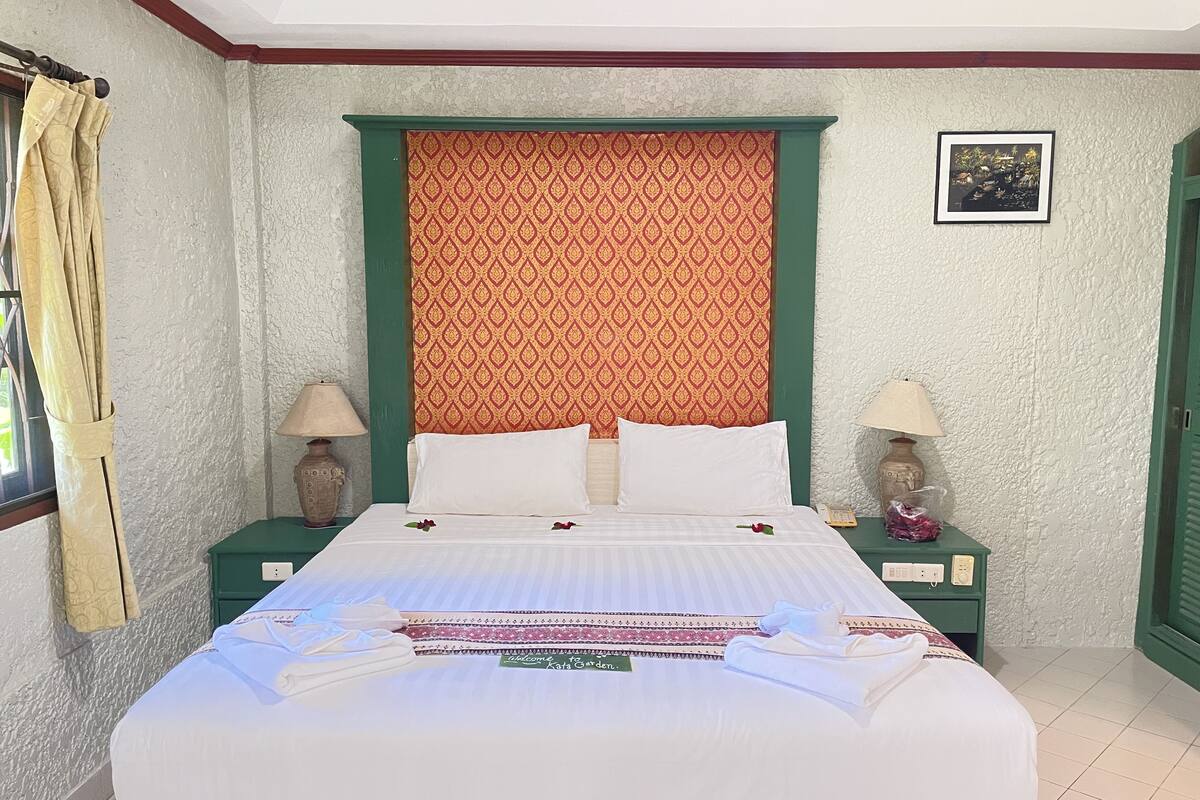 A neatly made bed with white linens and decorative pillows, a green headboard, and a patterned wall behind it. There are two bedside tables with lamps and a framed picture on the wall.
