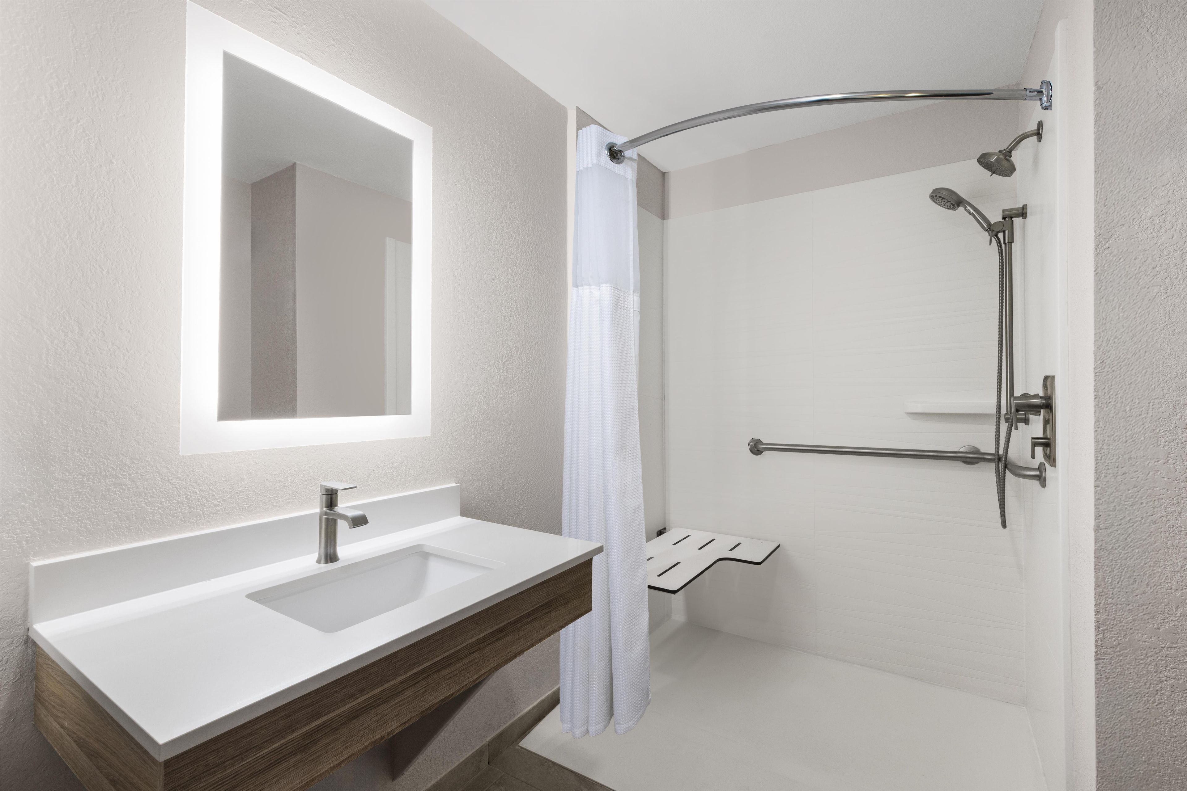 Un baño moderno con un lavamanos blanco, un espejo, una ducha con puerta de vidrio curvo y un mueble de tocador de madera.