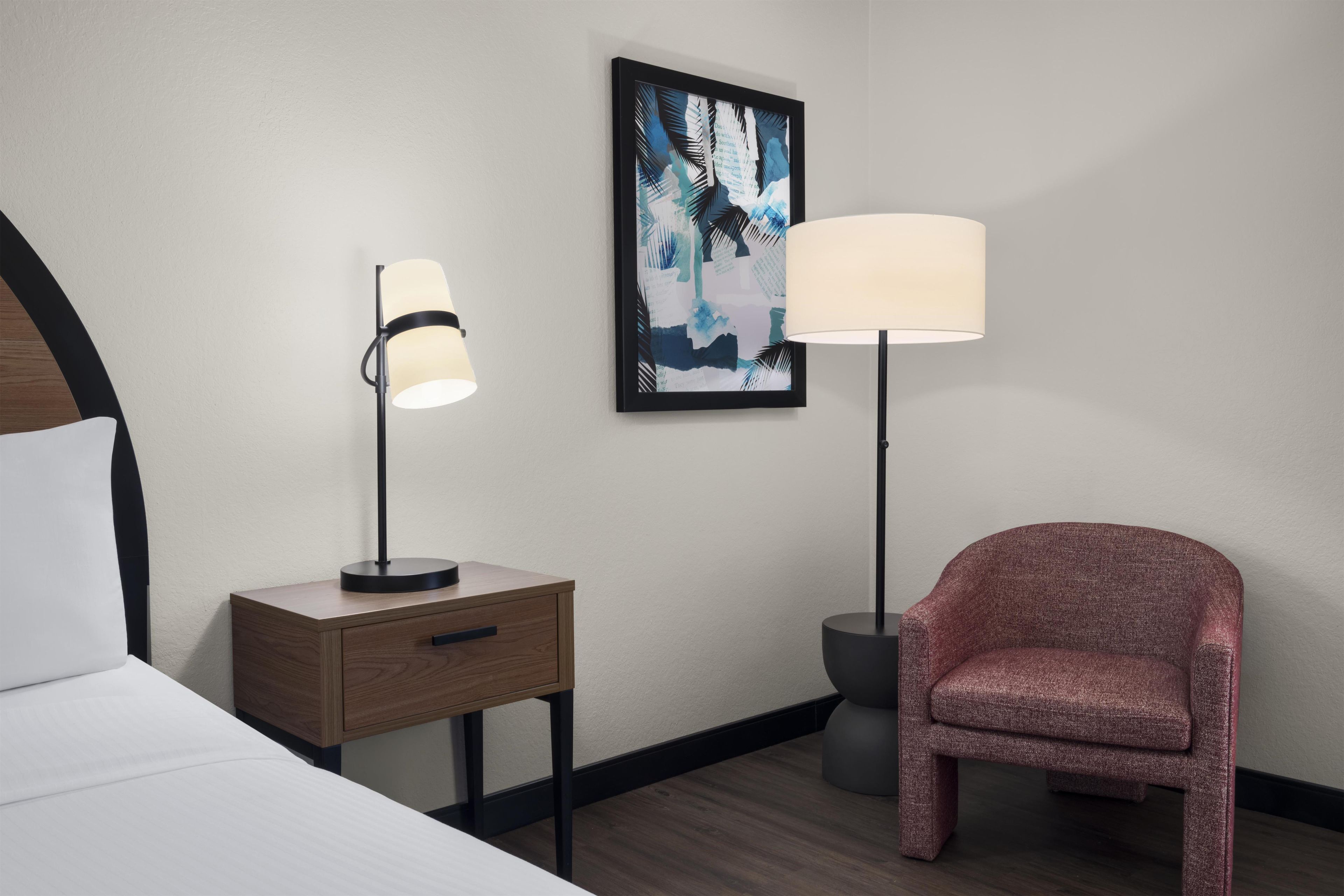 Habitación de hotel con una cama, una mesita de noche con lámpara, una silla y un cuadro abstracto enmarcado en la pared.