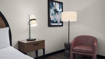Habitación de hotel con una cama, una mesita de noche con lámpara, una silla y un cuadro abstracto enmarcado en la pared.
