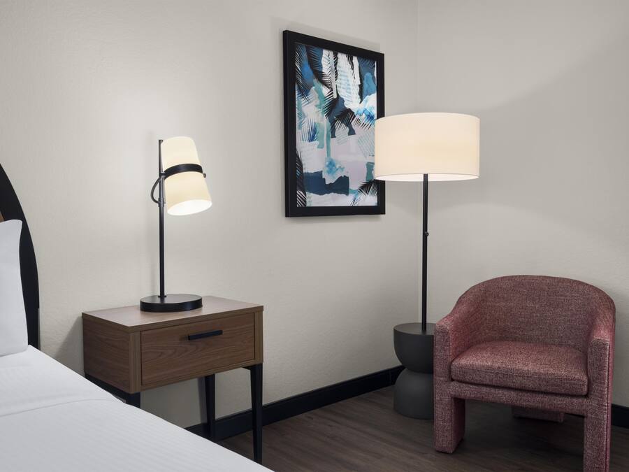 Habitación de hotel con una cama, una mesita de noche con lámpara, una silla y un cuadro abstracto enmarcado en la pared.