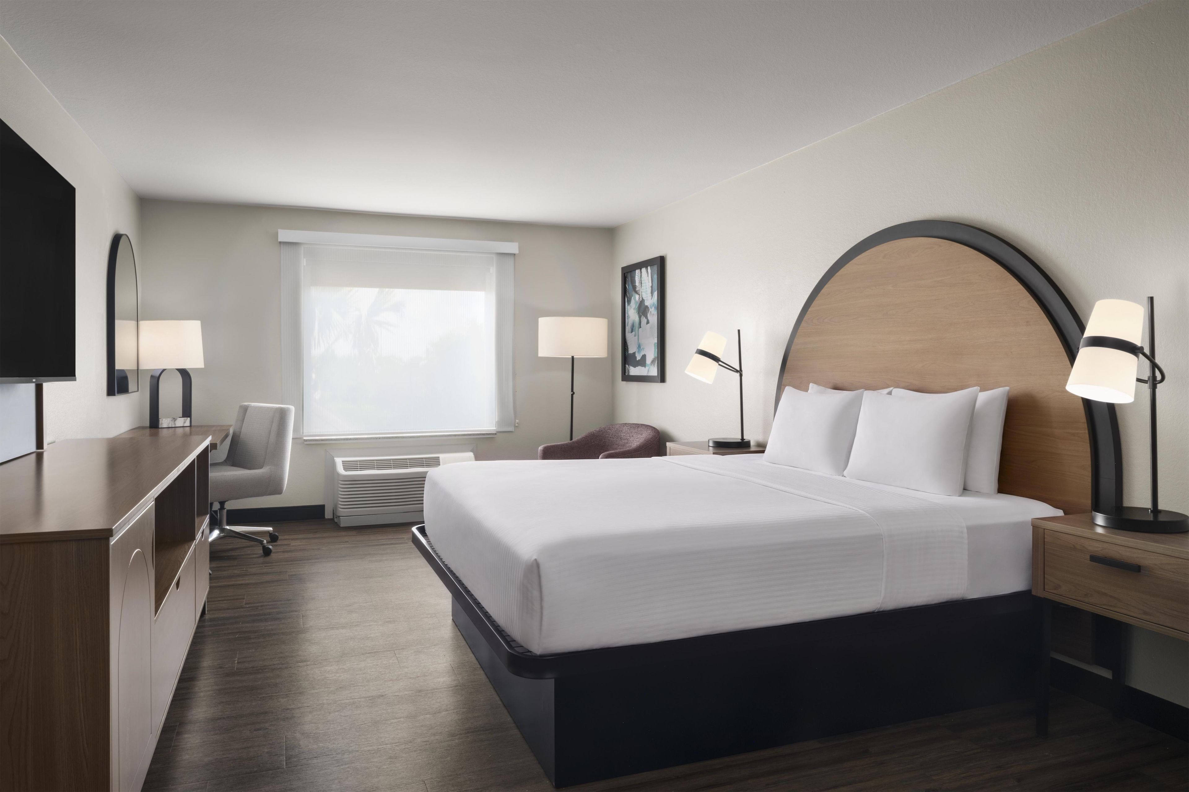 Una camera d'albergo con un letto grande, lampade ai comodini, una scrivania, una sedia, una televisione e una finestra con persiane.